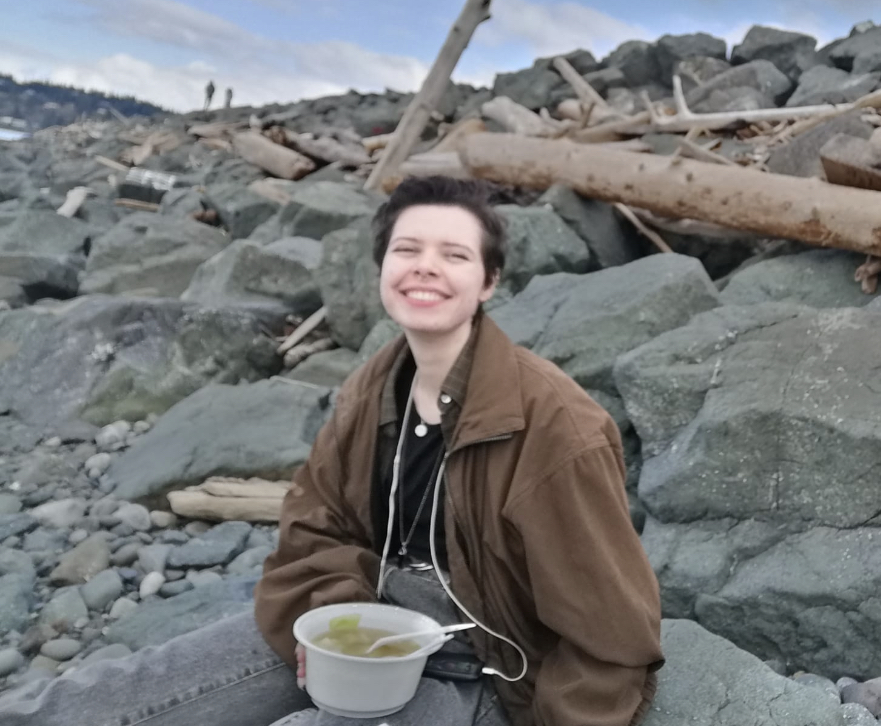 A photo of Juno sitting on some rocks at the beach. He has on a big brown jacket, grey jeans, and a black shirt. His hair is a growing-out buzzcut and they're smiling at the camera. A large white bowl of soup is sitting in their lap.